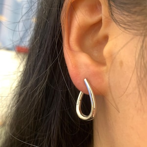 Pode incluir: Um close-up de um brinco prateado em forma de gota usado numa orelha. O brinco tem uma superfície lisa e polida e um design curvo. O brinco é uma peça de joalharia simples e elegante.