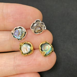 Puede incluir: Cuatro pequeños cabujones de concha de abulón engastados en biseles de metal plateado y dorado. Las conchas de abulón muestran una gama de colores, incluyendo azul, verde y púrpura. Los biseles tienen una forma ondulada y orgánica.