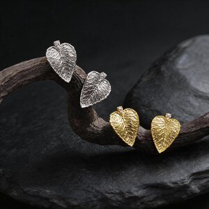 Peut inclure: Quatre boucles d'oreilles en forme de feuille. Deux sont argentées et deux sont dorées. Chaque boucle d'oreille présente un motif de veines de feuille détaillé et une petite gemme transparente en haut. Les boucles d'oreilles sont présentées sur une surface sombre et texturée.