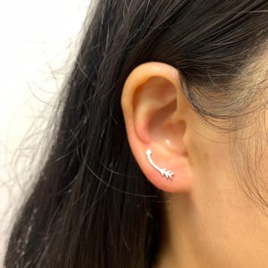 Może przedstawiać: Srebrny kolczyk wspinaczkowy jest noszony na uchu. Kolczyk zakrzywia się ku górze i ma mały wzór liścia na końcu. Kolczyk jest w minimalistycznym stylu.