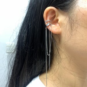 Puede incluir: Puño de oreja plateado con cadenas colgantes. El puño de oreja presenta un diseño curvo con una fila de pequeñas piedras transparentes en la banda superior. Tres delicadas cadenas cuelgan, creando un efecto en capas. La joya se lleva en la oreja.