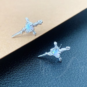 Puede incluir: Un par de pendientes de botón plateados. Cada pendiente presenta un diseño de espada con una piedra central ovalada e iridiscente. Los pendientes están colocados sobre un fondo marrón y negro.