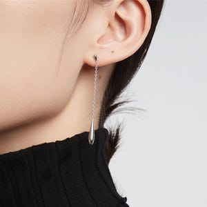 Puede incluir: Pendientes de gota de plata con una cadena delicada. Los pendientes se muestran en una modelo con cabello oscuro y un cuello de tortuga negro.