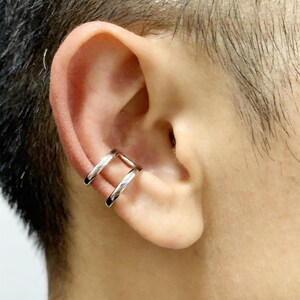 May include: A silver-colored ear cuff is worn on the ear. The cuff has a double-band design, with two parallel bars that wrap around the ear cartilage. The ear cuff is a simple, modern accessory.