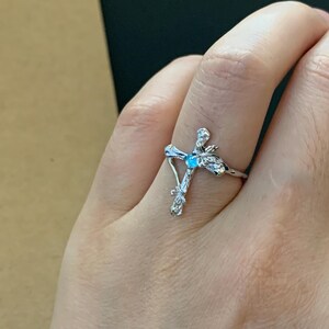 May include: A silver ring with a cross design featuring a blue gemstone. The cross is detailed with intricate branches and leaves.