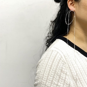 May include: A pair of silver-toned, long dangle earrings. The earrings feature a looped design that connects to a long, thin chain that hangs down. The earrings are worn by a person with dark hair and a white sweater.