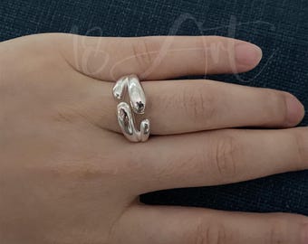 May include: A silver ring with a distinctive, fluid design. The ring has a wrap-around style, featuring two curved, rounded elements. The polished metal reflects light, emphasizing the ring's smooth surface and contemporary look.