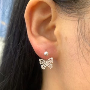 May include: A pair of silver butterfly earrings. The earrings feature a small, round stud at the top and a delicate, openwork butterfly design with small, sparkling accents that hangs below. The earrings are worn on an ear.