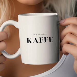 May include: White ceramic coffee mug with the text "MEN FØRST... KAFFE" in black. The mug has a curved handle and is held by a person. The mug is a standard size for coffee or tea.