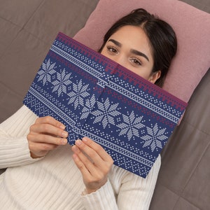 Pu&ograve; includere: Una donna &egrave; sdraiata su un letto con un cuscino rosa dietro la testa. Sta tenendo un libro blu e bianco con un motivo a fiocco di neve. Il libro le copre il viso.