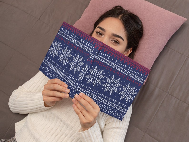 Pu&ograve; includere: Una donna &egrave; sdraiata su un letto con un cuscino rosa dietro la testa. Sta tenendo un libro blu e bianco con un motivo a fiocco di neve. Il libro le copre il viso.