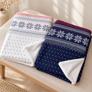 Könnte beinhalten: Zwei gefaltete Decken mit einem nordisch inspirierten Muster aus Schneeflocken und kleinen Punkten. Eine Decke ist weiß mit marineblauen Details und rotem Rand, die andere ist marineblau mit weißen Details und einem dunkelroten Rand, beide mit weißem Fleece.