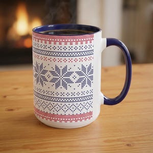 Könnte beinhalten: Ein weißer Keramikbecher mit dunkelblauem Innenraum und Henkel, verziert mit einem sich wiederholenden rot-blauen nordischen Schneeflocken- und geometrischen Muster auf der Außenseite, gefüllt mit einem dunklen Getränk.