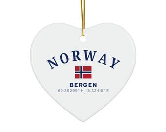 May include: A heart-shaped white ceramic ornament with the word "NORWAY" in navy blue arched across the top. Below is the Norwegian flag and the city name "BERGEN". Coordinates are printed at the bottom. A gold string is attached for hanging.