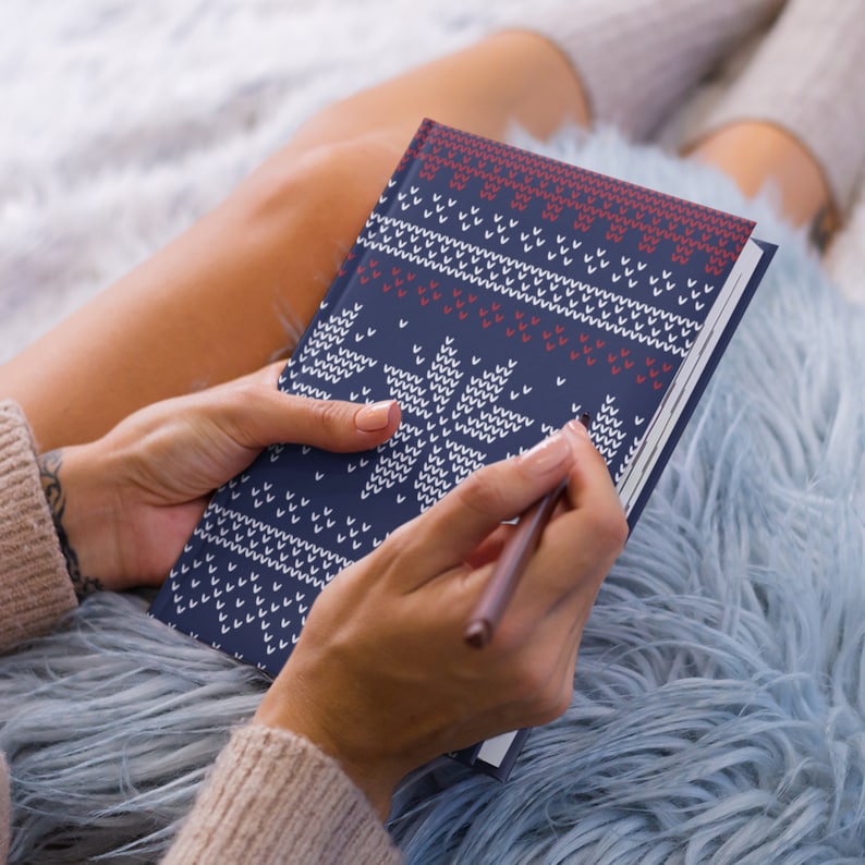 Pu&ograve; includere: Un quaderno blu con un motivo a fiocco di neve bianco e rosso. Il quaderno &egrave; aperto e una persona sta scrivendo al suo interno con una matita.