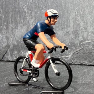 May include: A miniature figurine of a cyclist wearing a white, red, and black helmet and a dark blue jersey. The cyclist is positioned on a white and red racing bicycle with black wheels, pedals, and handlebars, captured in a forward-leaning pose.