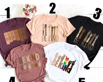 Può includere: Cinque magliette diverse con la frase "Be Kind" e una grafica di mani in diverse tonalità di pelle. Le magliette sono di vari colori, tra cui il granata, il pesca, il nero e il rosa.