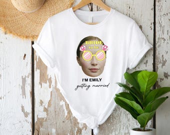 May include: White t-shirt with a graphic of a woman wearing novelty glasses and the text "Bride to be". Below the graphic is the text "I'm Emily getting married". A straw hat hangs in the background.
