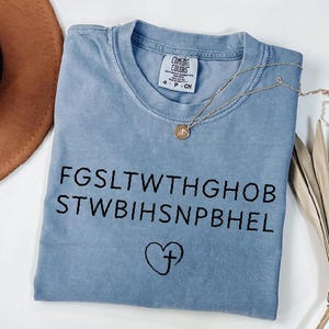 May include: A light blue t-shirt with black text and a heart with a cross graphic. A gold necklace with a pendant is draped across the shirt. The shirt has a Comfort Colors tag.