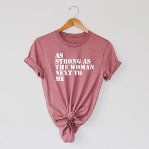 Pode incluir: Uma camiseta cor-de-rosa com o texto "AS STRONG AS THE WOMAN NEXT TO ME" impresso em branco.