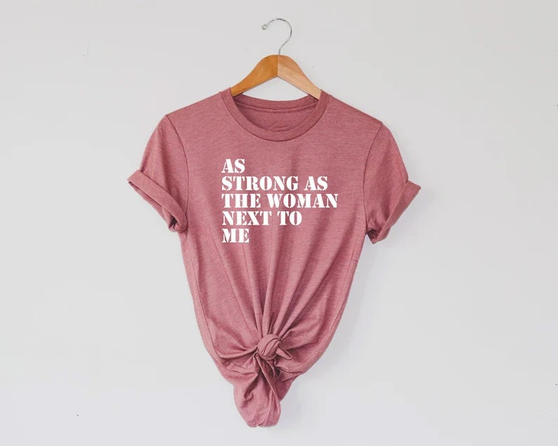 Pode incluir: Uma camiseta cor-de-rosa com o texto "AS STRONG AS THE WOMAN NEXT TO ME" impresso em branco.