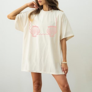 May include: Cream-colored t-shirt with a pink seashell design and text. The text reads "Bailey's Bachelorette Hawaii May 2023". The shirt has short sleeves and a relaxed fit, suitable for casual wear.