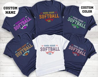 May include: Assortment of navy blue and white softball t-shirts. Each shirt features the text "YOUR NAME SOFTBALL 10" in various colors, with a softball graphic. The shirts are displayed on a light wood surface.