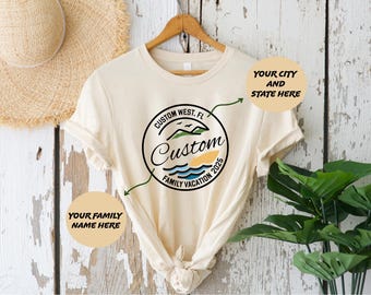 May include: A beige t-shirt with a custom family vacation logo. The logo is a circle with a beach scene, palm trees, and the text "Custom Family Vacation 2025". There are two text boxes on the image, one says "Your Family Name Here" and the other says "Your City and State Here".