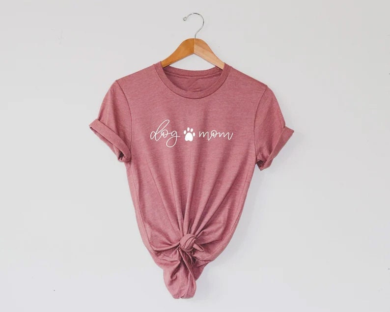 Puede incluir: Una camiseta de color malva con el texto "dog mom" en letras blancas con una huella de pata entre las palabras.