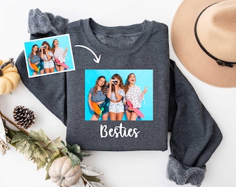 May include: Dark gray sweatshirt with a photo of three women on the front, with the word "Besties" in white script. A smaller photo of the same women is above the larger photo. A tan hat is on the right.