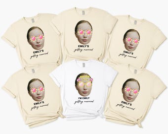 May include: A collection of cream-colored and white t-shirts. Each shirt features a graphic of a woman's face wearing sunglasses with a floral design. The text "EMILY'S getting married" is printed on most shirts. One shirt says "I'M EMILY getting married".