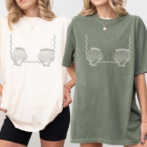 May include: Ivory and moss green t-shirts with a seashell graphic. The graphic includes the text "Bailey's Bachelorette Hawaii July 2023". The shirts are oversized and have short sleeves. The ivory shirt is paired with black shorts.