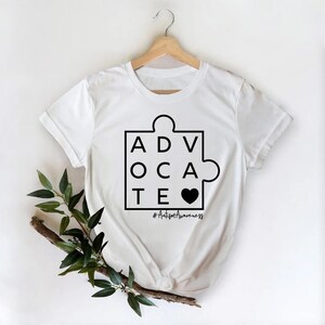May include: White t-shirt with a black puzzle piece design that spells out "ADVOCATE" with a black heart in the middle. The text "#AutismAwareness" is below the puzzle piece.