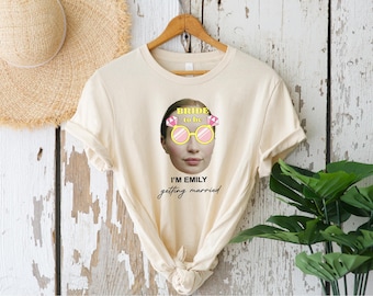 May include: Cream-colored t-shirt with a graphic of a woman's face wearing sunglasses and a tiara that says "Bride to be." The text "I'm Emily getting married" is below the graphic. The shirt is hanging on a wooden hanger.