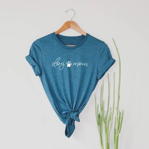 Puede incluir: Una camiseta azul turquesa con el texto "dog mom" en letras blancas con un s&iacute;mbolo de huella de pata entre las palabras.