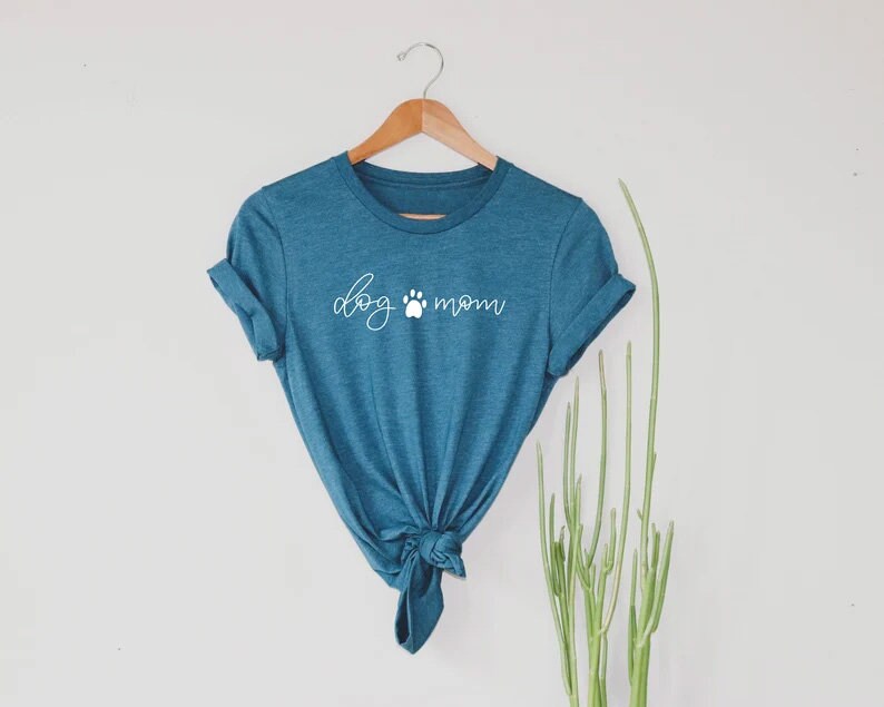 Puede incluir: Una camiseta azul turquesa con el texto "dog mom" en letras blancas con un s&iacute;mbolo de huella de pata entre las palabras.
