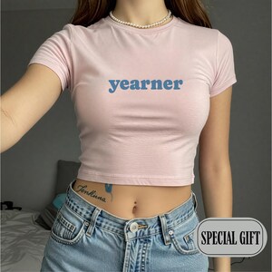 May include: A light pink short-sleeved crop top featuring the word "yearner" printed in blue letters across the chest. The garment has a crew neckline and a fitted silhouette, paired with blue denim jeans that have a visible button and zipper.