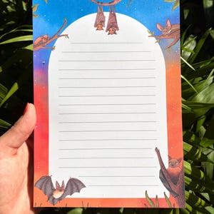 May include: A lined notepad with a bat theme. The notepad features illustrations of bats hanging from branches and flying against a gradient background of blue, orange, and red. The notepad is held by a hand.