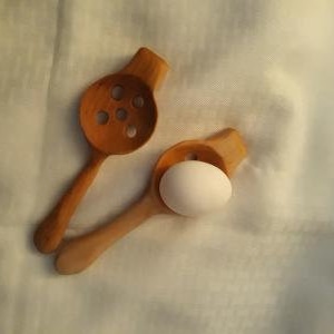 May include: Two wooden cooking spoons, one with holes, and a white egg. The spoons are made of light brown wood and are positioned on a white, textured surface. The egg rests in one of the spoons.