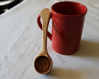 May include: A wooden spoon with a rounded bowl and a long handle. The spoon is resting on a white tablecloth next to a red ceramic mug.