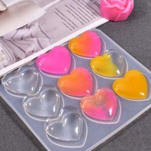 Può includere: Uno stampo in silicone con otto cavità a forma di cuore. Lo stampo è riempito con cuori di resina colorati in tonalità di rosa, arancio e giallo.