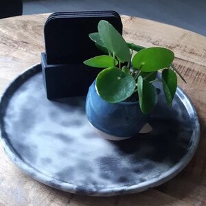 Pu&ograve; includere: Un vassoio rotondo grigio e bianco con un porta sottobicchieri nero e una piccola pianta verde in un vaso blu.