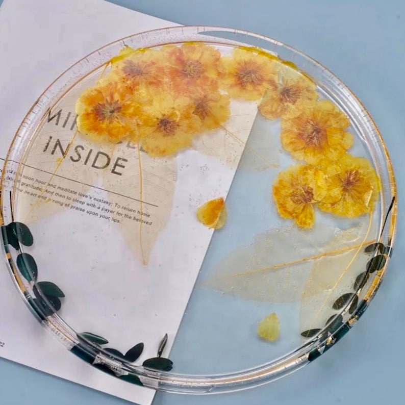 Pu&ograve; includere: Un vassoio rotondo in resina trasparente con un bordo dorato e un design a foglie nere e dorate. Il vassoio &egrave; pieno di fiori gialli pressati.
