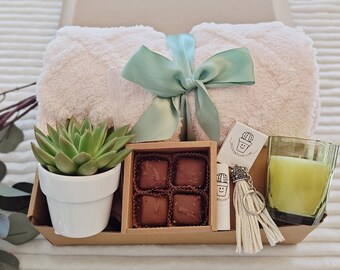 May include: A gift basket with a white fluffy blanket, a small potted succulent, a box of four chocolate squares, a candle in a clear glass jar, and a keychain with a cactus charm and a white tassel.