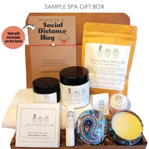 Op de afbeelding: Een cadeaudoos gevuld met spa-producten, waaronder badzout, suikerscrub, zeep, lippenbalsem, badbom en kaars. De doos is versierd met bruin en wit gepatroneerd papier en heeft een kaart met de tekst "Social Distance Hug".