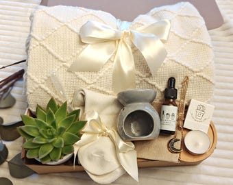 May include: A gift basket filled with self-care items, including a white knitted blanket, a gray ceramic oil diffuser, a bottle of essential oil, a pair of white socks, a succulent plant, a candle, and a gold necklace with a crescent moon pendant.
