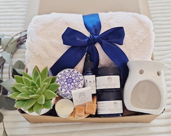 May include: A gift basket filled with self-care items, including a white towel, a succulent plant, a blue and white patterned candle, a white ceramic wax warmer, and two blue jars of body butter. The basket is tied with a blue ribbon.