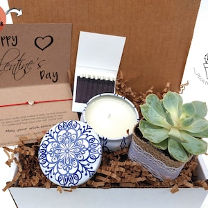 May include: A white gift box filled with a variety of items, including a small succulent plant, a candle in a blue and white tin, a matchbook, and a red string bracelet. The box is decorated with brown paper shreds and a card that reads "Happy Valentine's Day".