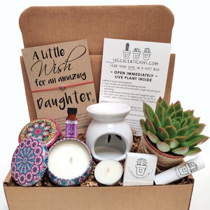 May include: A brown cardboard box filled with a gift set for a daughter. The box contains a small succulent plant, a white ceramic oil diffuser, a candle, a small tea light, a lip balm, and a small bottle of essential oil. The box also includes a card with the text "A Little Wish for an Amazing Daughter".