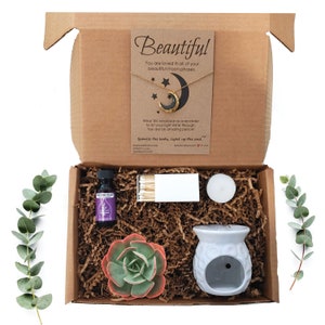 May include: A gift box filled with self-care items, including a crescent moon necklace, essential oil, matches, a candle, a succulent plant, and a white ceramic oil diffuser. The box is decorated with brown paper shreds and a card that reads "Beautiful. You are loved in all of your beautiful moon phases. Wear this necklace as a reminder to let your light shine through. You are an amazing person! Sparkle the body, light up the soul."